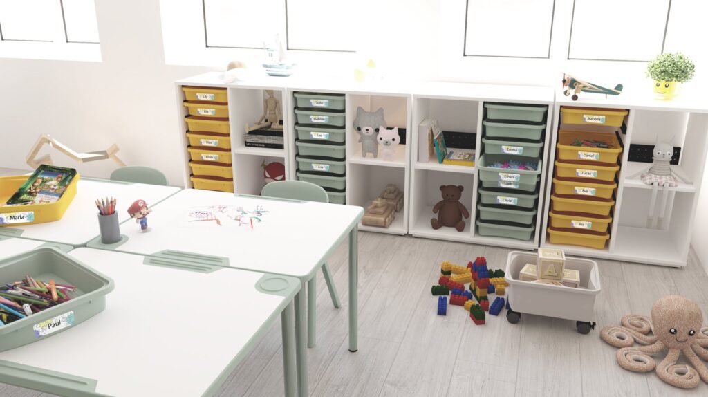 Sala com estantes brancas, recipientes amarelos e verdes, brinquedos e mesas com desenhos e lápis de cor.