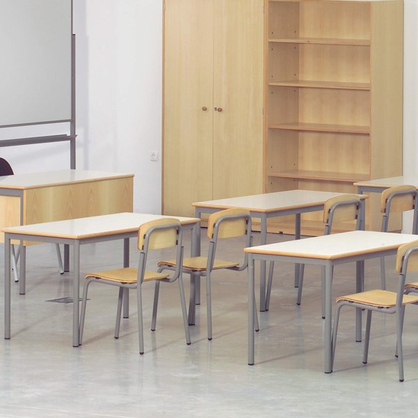 Sala de aula com mesas e cadeiras de madeira, mesa de professor, quadro branco, armário e estante.