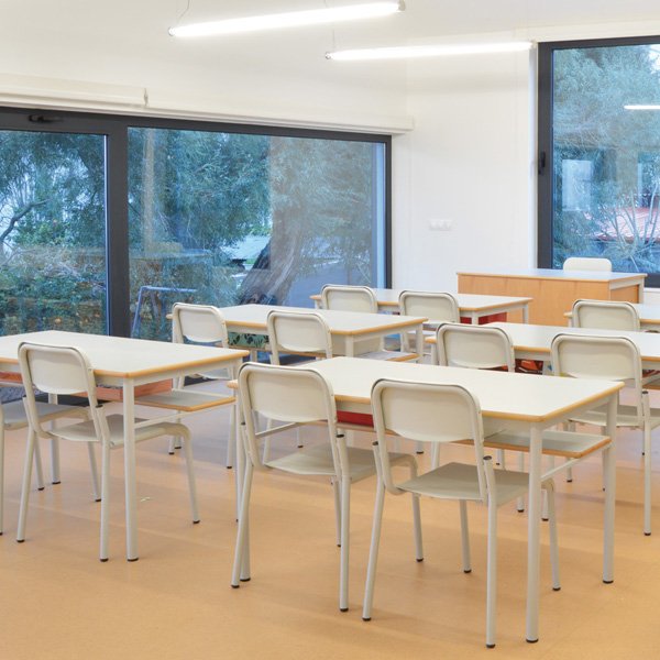 Sala de aula com mesas e cadeiras em filas, mesa de professor e grandes janelas.