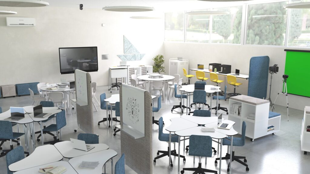 Sala de aula com mesas brancas agrupadas, cadeiras azuis e amarelas, computadores, fundo verde e câmara em tripé.