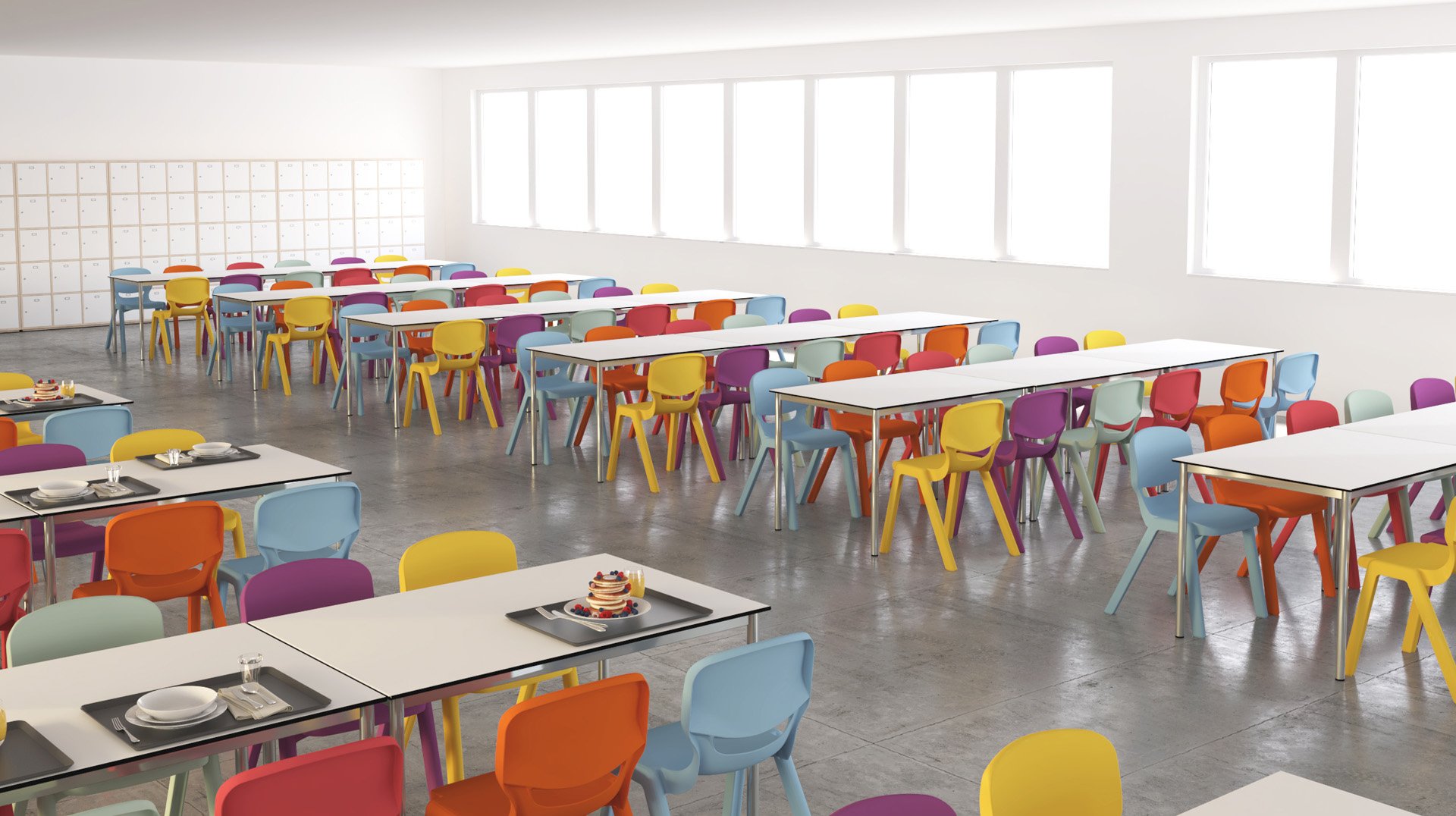 Una cafetería muy iluminada con sillas de colores en naranja, amarillo, morado y rojo alrededor de mesas blancas. En algunas mesas hay bandejas con comida y en la pared del fondo hay taquillas. Grandes ventanales dejan entrar la luz natural.