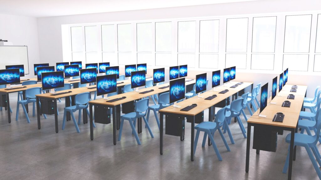 Sala de informática com mesas em filas, monitores, teclados, ratos e cadeiras azuis.