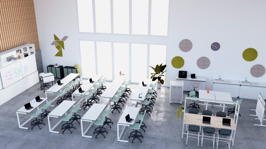 Sala com mesas em filas, computadores portáteis, cadeiras verdes e zona de trabalho com bancos altos.