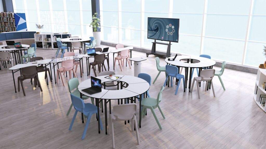 Sala com mesas redondas, cadeiras de várias cores, computadores portáteis, ecrã e estantes.