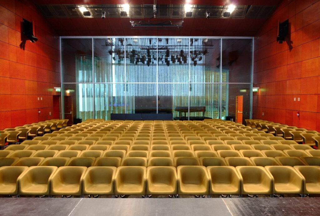 Auditório com filas de cadeiras bege diante de um palco com piano de cauda.