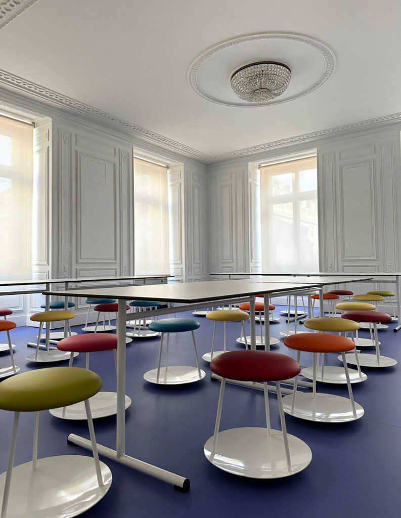 Sala com paredes ornamentadas, lustre, mesas e bancos redondos de várias cores sobre pavimento azul.