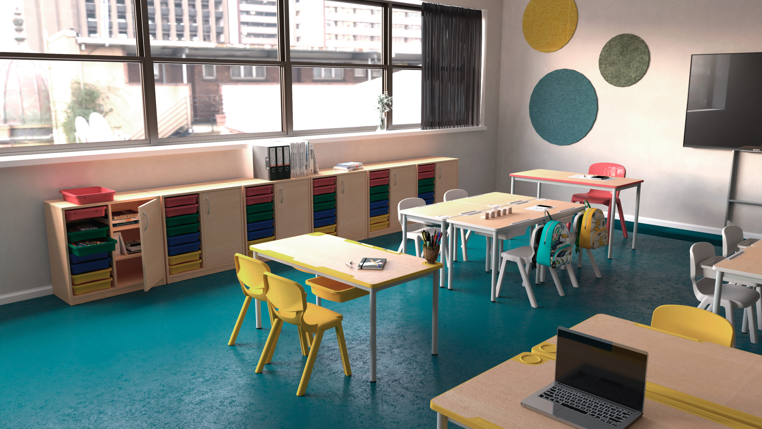 Sala de aula com mesas e cadeiras de várias cores, móveis de arrumação e computador portátil.
